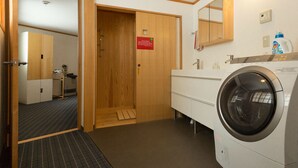 A hotel bathroom with a wooden door, a washing machine, a sink, a mirror, and a red sign on the wall. - Nozawa Cottage (Nozawaonsen)