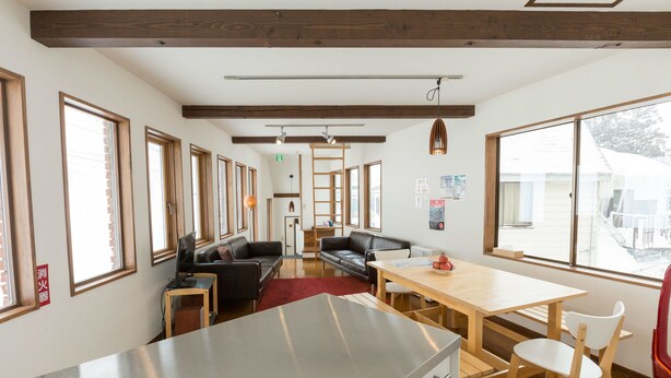 A modern kitchen with a stainless steel countertop, wooden cabinets, and a red rug. There is a wooden dining table with chairs and a black leather couch in the background. - Nozawa Cottage (Nozawaonsen)