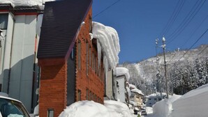 A snowy street with a brick building, a white car, and a person in green. - Nozawa Cottage (Nozawaonsen)