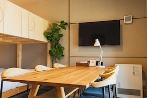A room with a wooden table, chairs, a television mounted on the wall, a plant, and a small white table with a lamp. - The Base Sapporo Susukino (Sapporo)