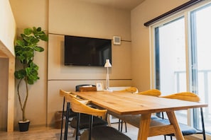 A modern room with a wooden dining table, chairs, a potted plant, a flat-screen TV mounted on the wall, and a window with a view. - The Base Sapporo Susukino (Sapporo)