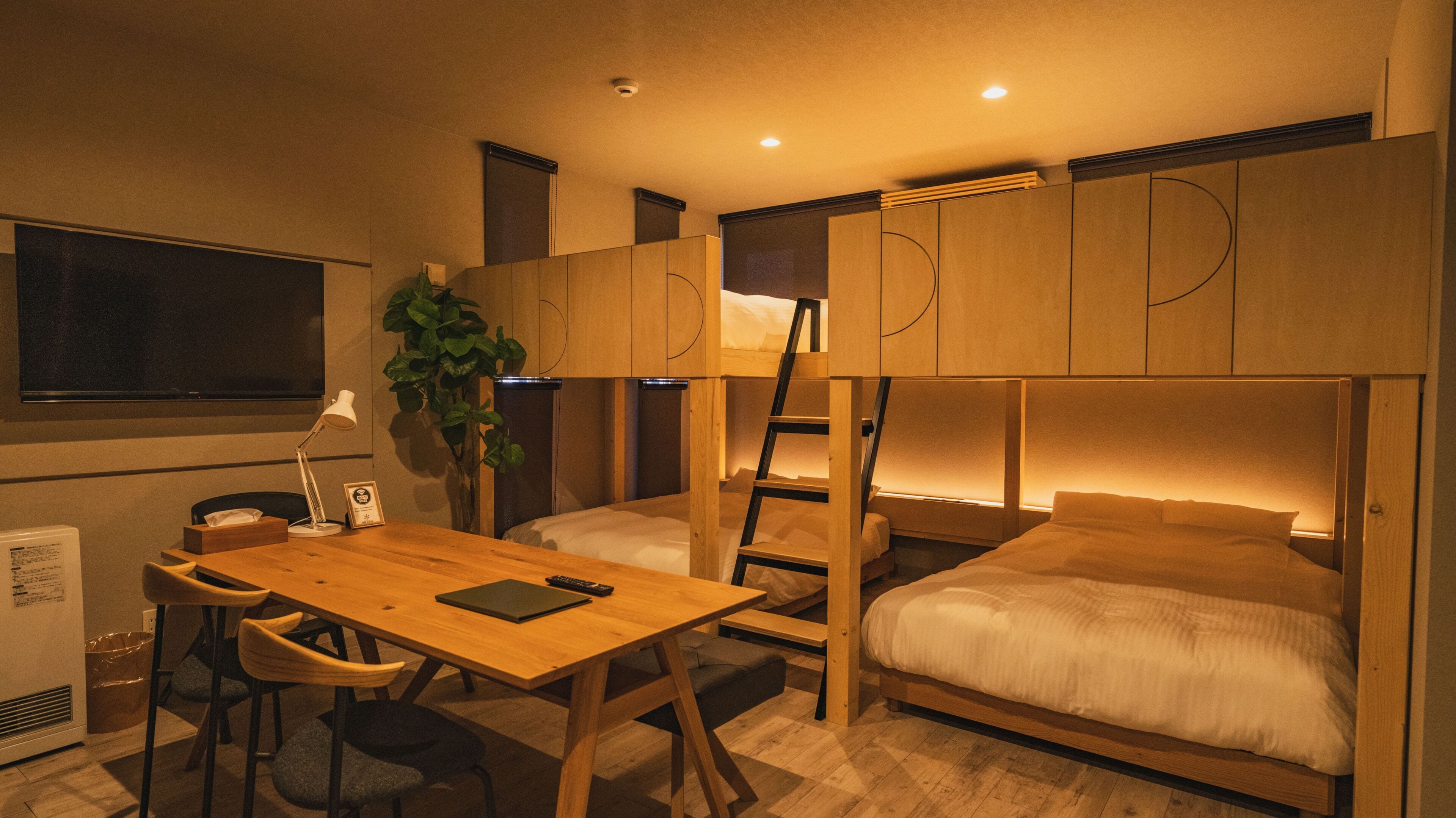 Een kleine kamer met stapelbedden, een bureau, stoelen en een plant.