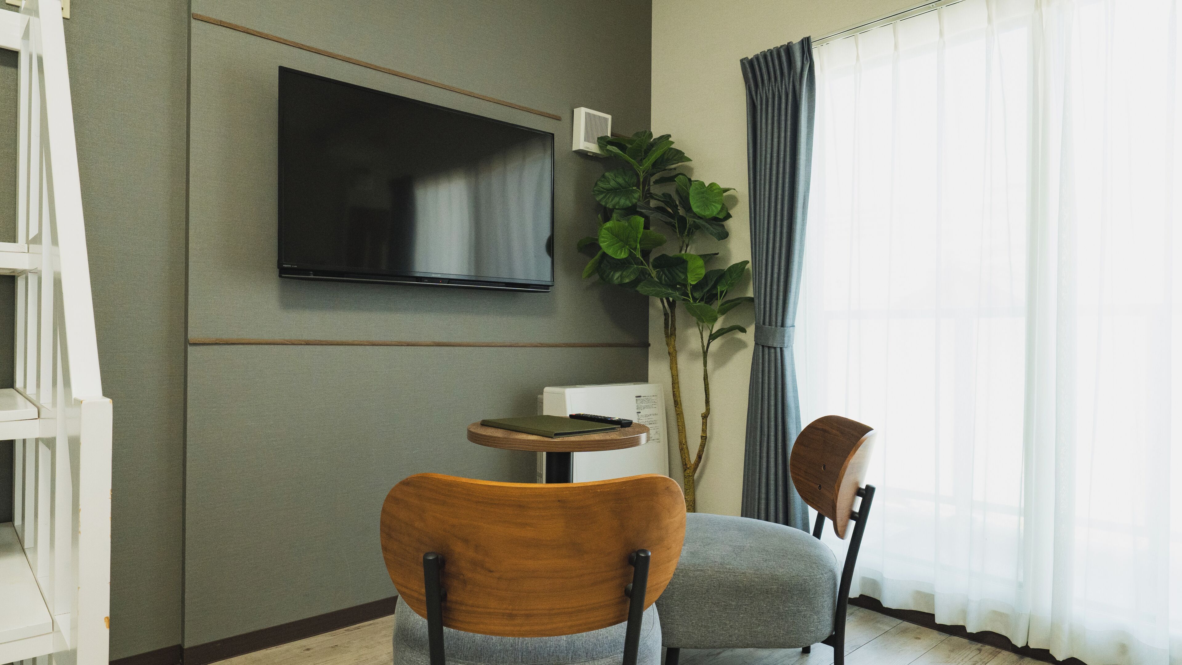 Een moderne kamer met een flatscreen televisie aan de muur, een klein rond tafeltje met een boek en afstandsbediening, twee stoelen, een potplant en een raam met gordijnen.