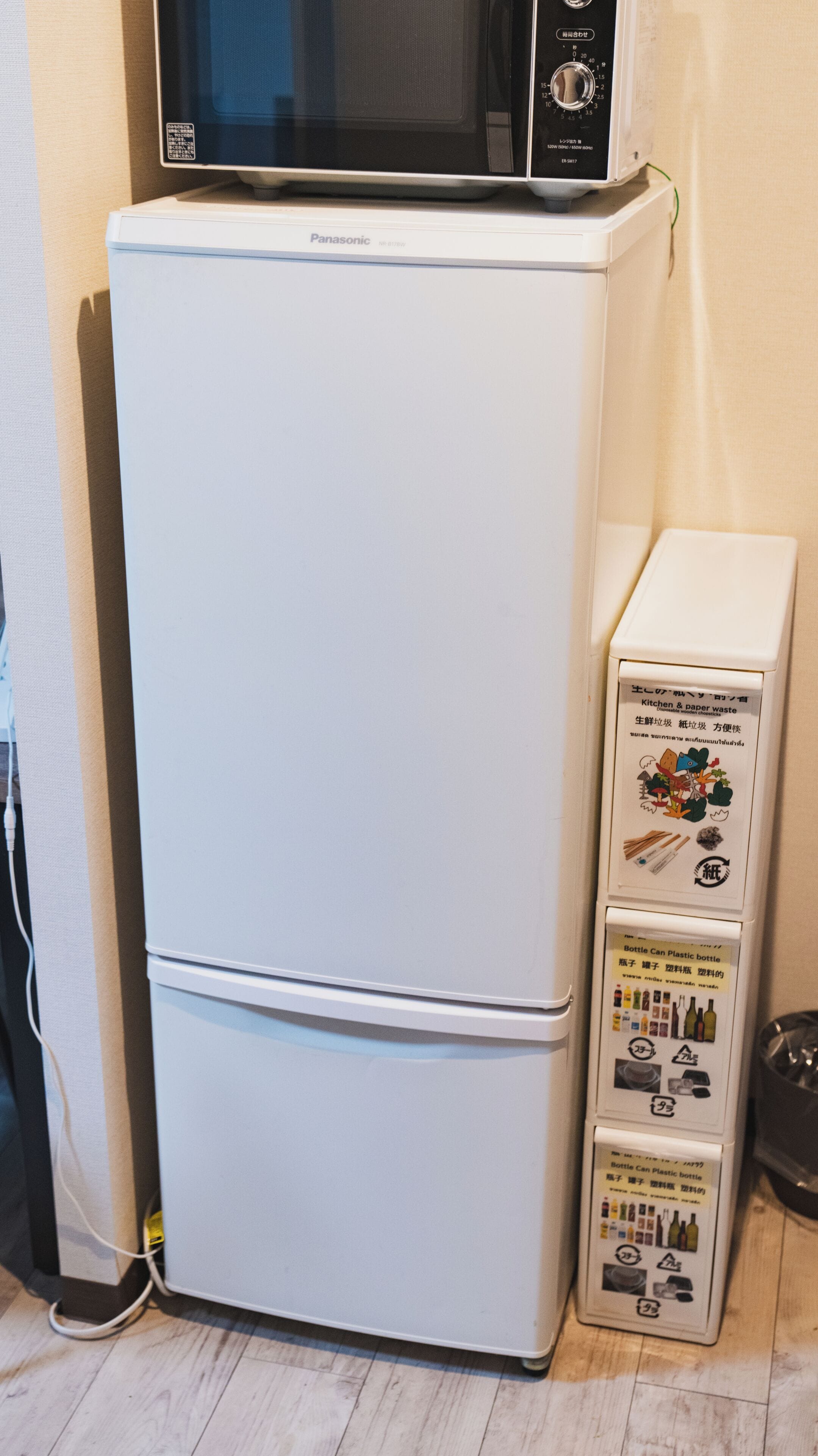 A white refrigerator with a microwave on top, a lower compartment, and a recycling bin with Japanese text.