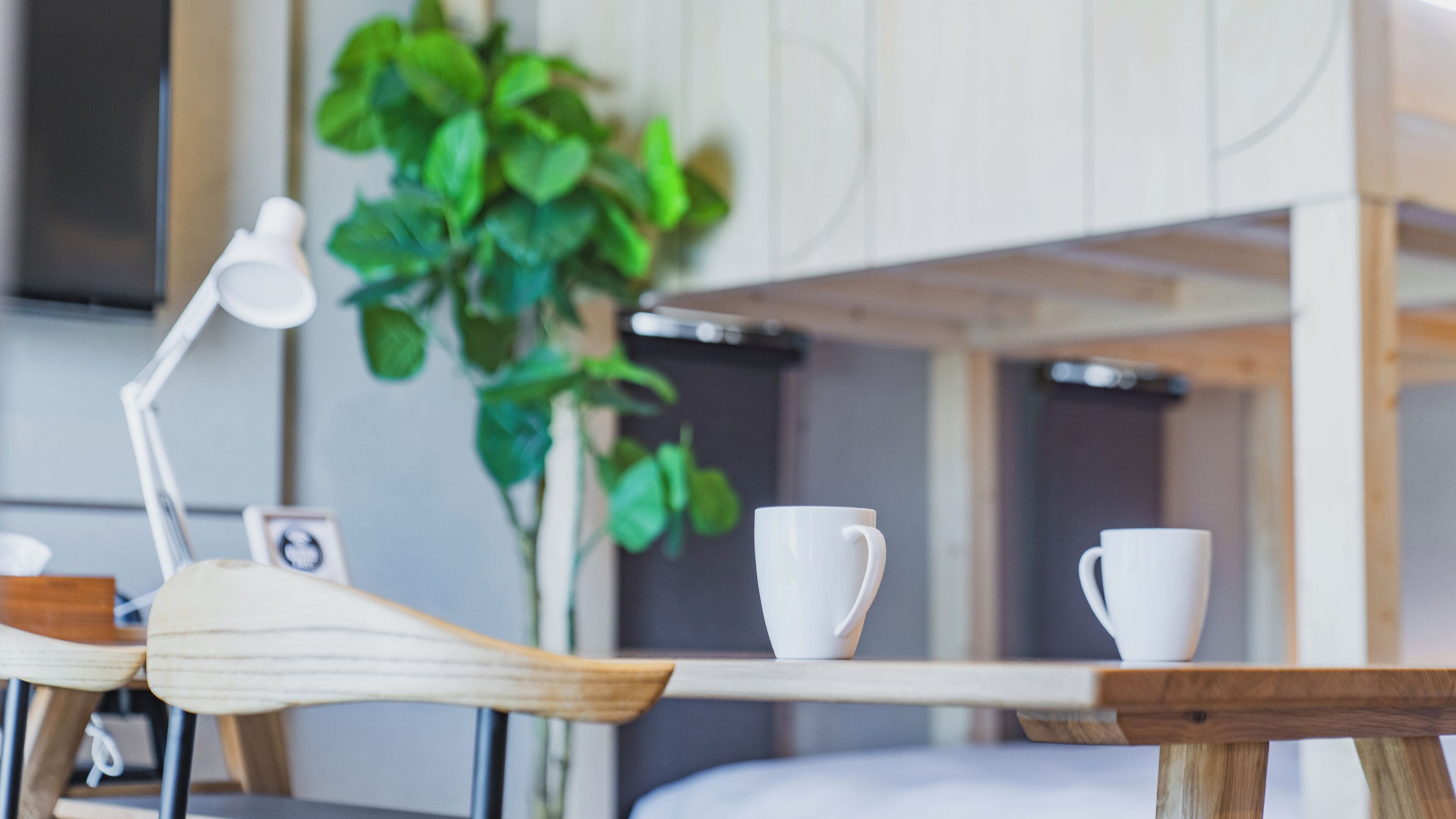 A wooden table with two white cups and a lamp, a plant, and a bed in the background.