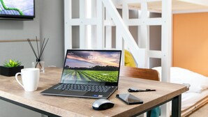 Un portátil sobre un escritorio de madera con una taza blanca, un teléfono inteligente y un par de auriculares. Una cama con ropa blanca se ve al fondo.