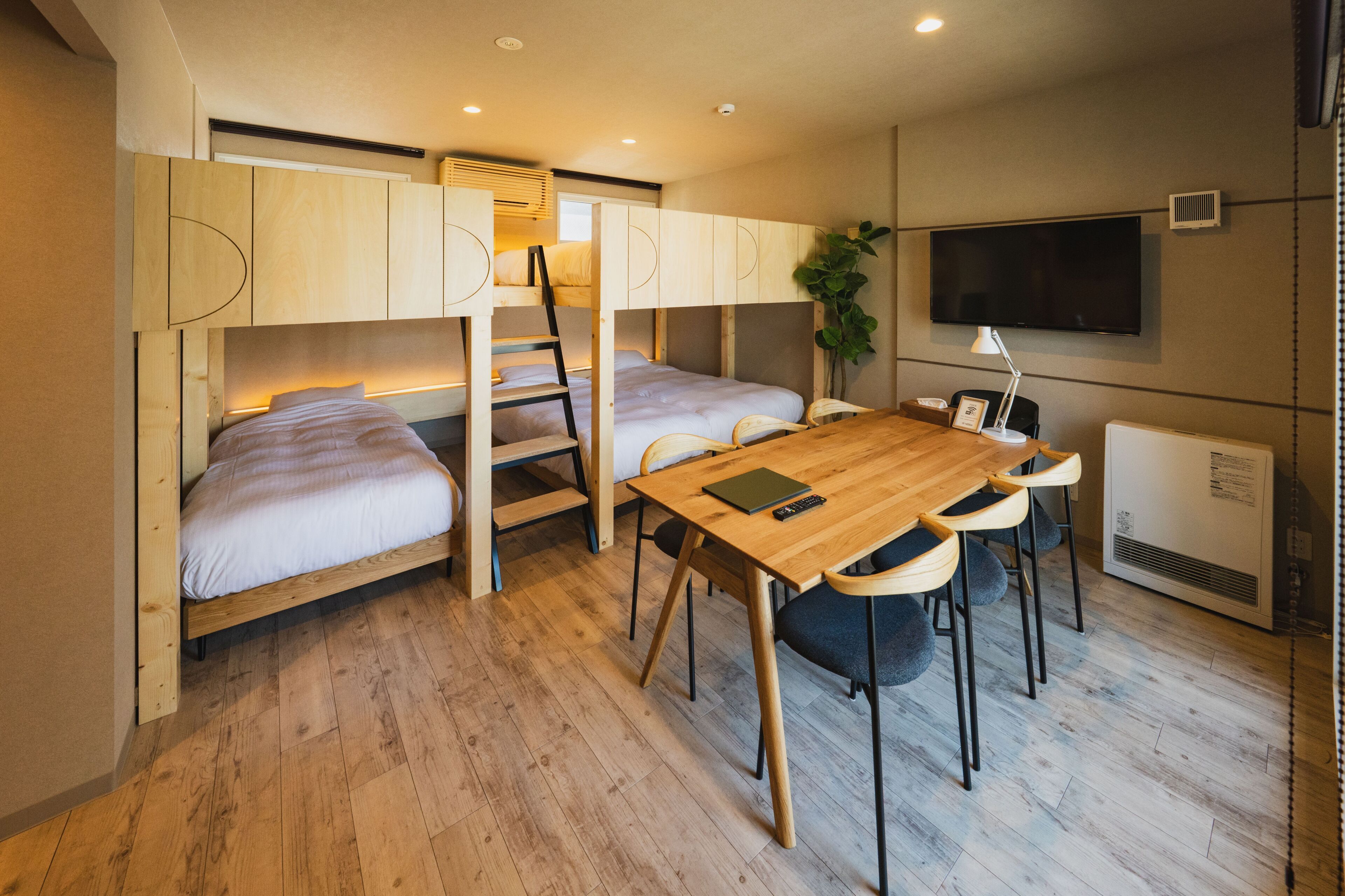 A compact room with bunk beds, a wooden table with chairs, a desk, a television, and a radiator.