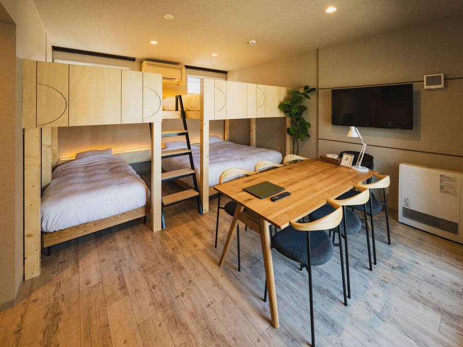 A compact room with bunk beds, a wooden table with chairs, a desk, a television, and a radiator.