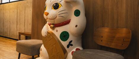 A large cat statue with a red collar holding a coin, placed between two chairs in a room with wooden flooring and a wooden wall.