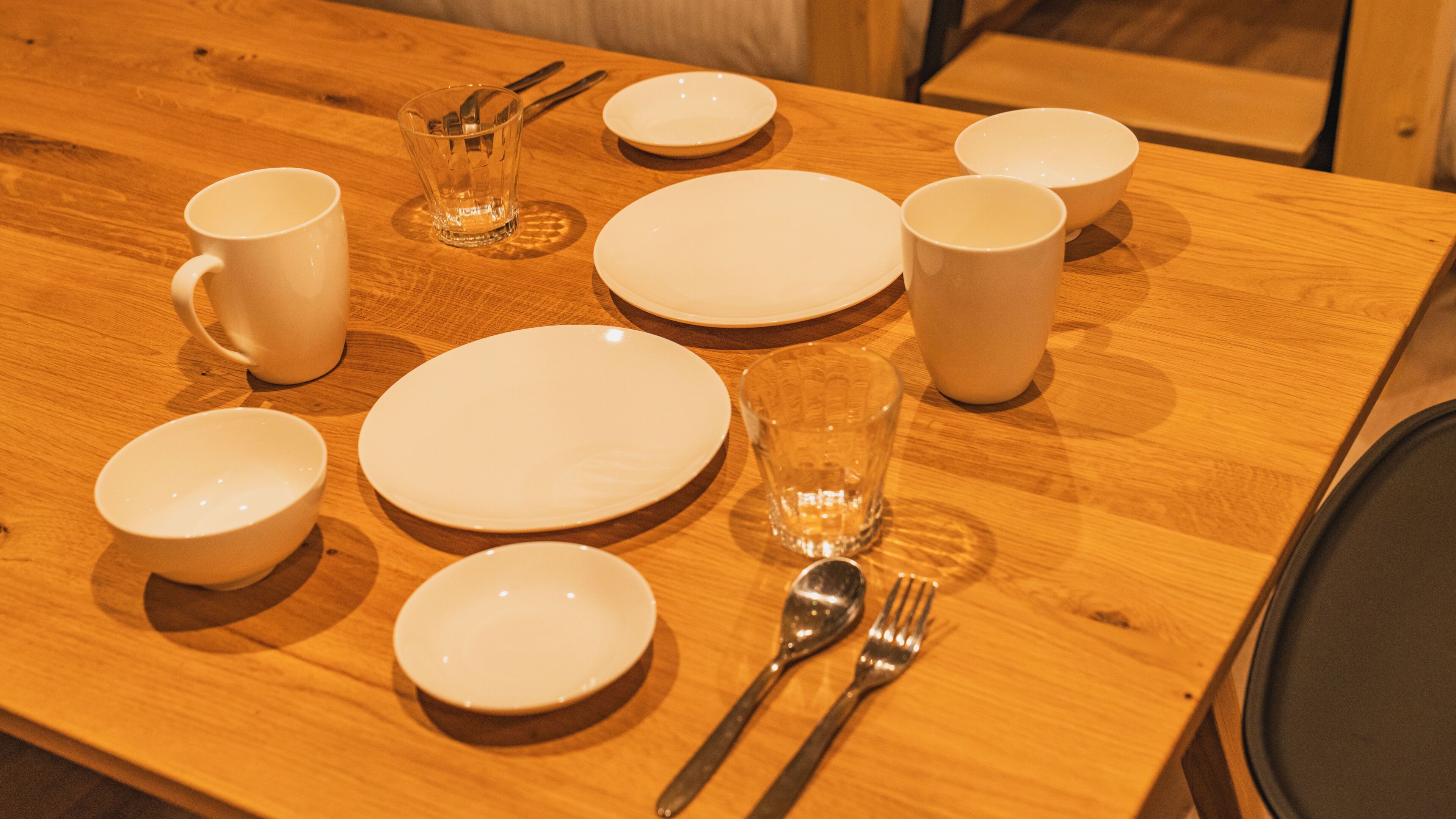 Une table de salle à manger en bois, mise avec des assiettes, des bols, des tasses et des couverts blancs.