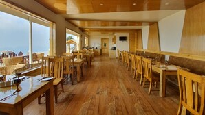 A spacious dining area with wooden tables and chairs, large windows offering a view of the sea, and a kitchen area in the background. - The Alpine Chalet Resort (Dhari)