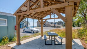 Ein hölzernes Pavillon mit Picknicktisch und Bänken, auf einer Betonplattform mit Blick auf eine Straße und Gebäude im Hintergrund.