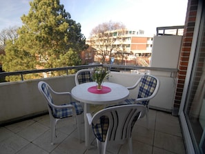 Uma varanda com uma mesa redonda e quatro cadeiras, um vaso com flores e vista para edifícios e árvores.