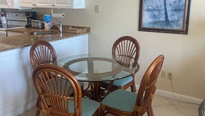 Un coin repas avec une table à plateau en verre entourée de chaises en osier dotées de coussins bleus, une cuisine avec des meubles blancs et un four à micro-ondes, et un tableau représentant un palmier accroché au mur.