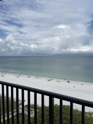 A beach with a railing in the foreground, a clear view of the ocean, and a cloudy sky.