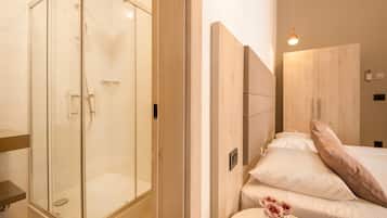 A modern bathroom with a glass-enclosed shower, a wooden vanity, and a bed with white linens and a pink pillow.