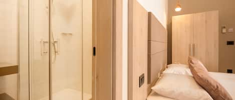 Un baño moderno con ducha acristalada, tocador de madera y cama con ropa de cama blanca y almohada rosa.