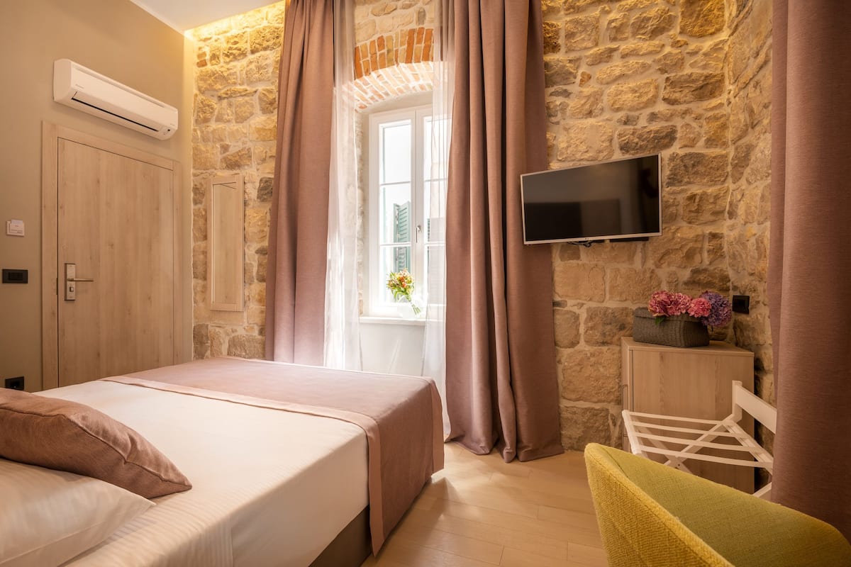 A bedroom with a bed, a TV mounted on a stone wall, a window with curtains, and a wooden cabinet.