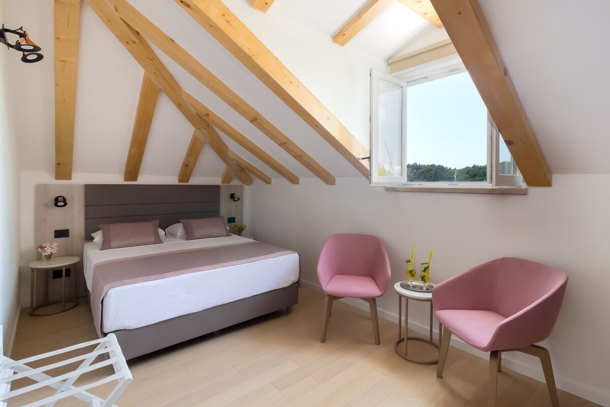 A bedroom with a bed, two pink chairs, a small table, and a window with a view.