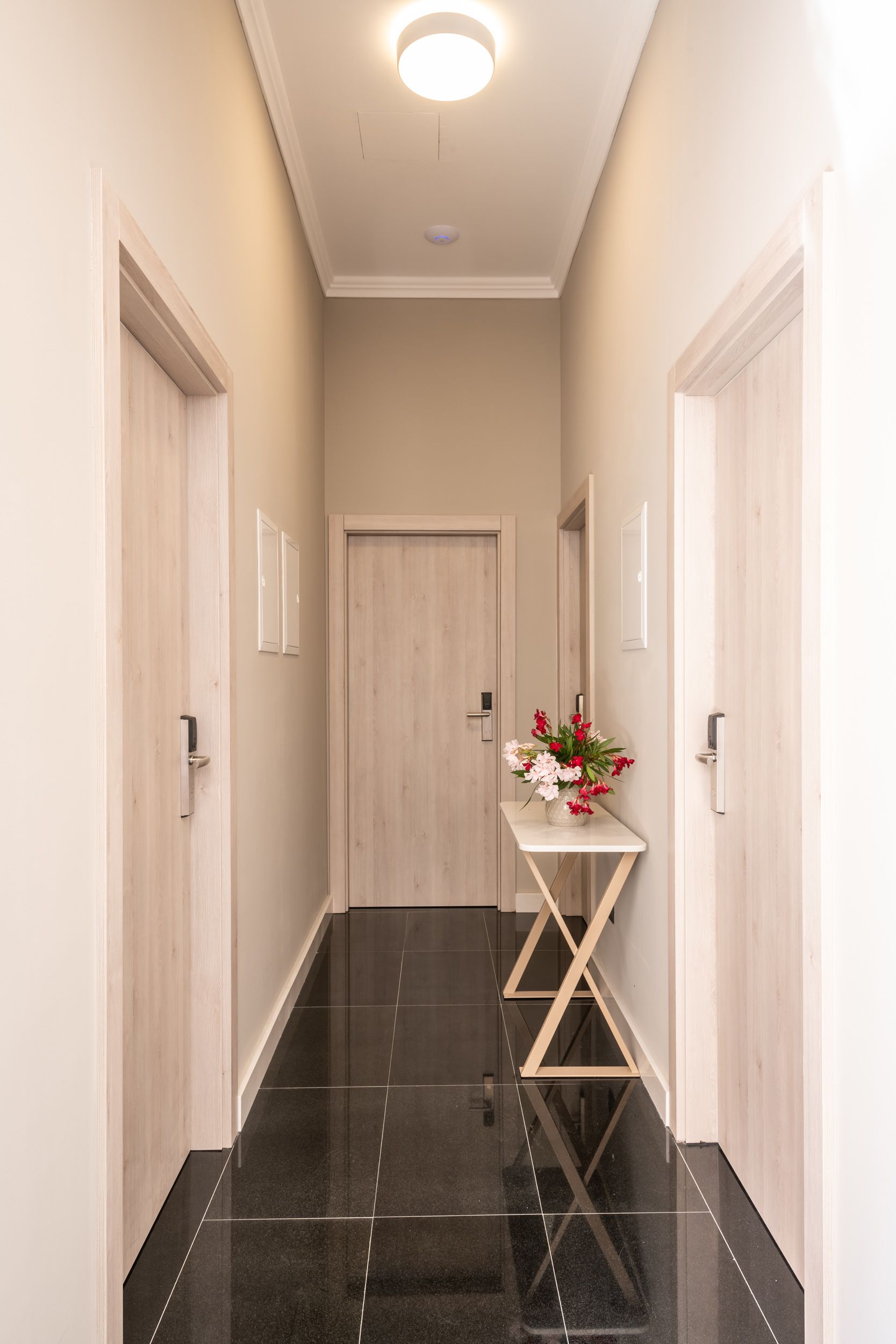 A hallway with a wooden door at the end, a small table with a vase of flowers, and a light fixture on the ceiling.