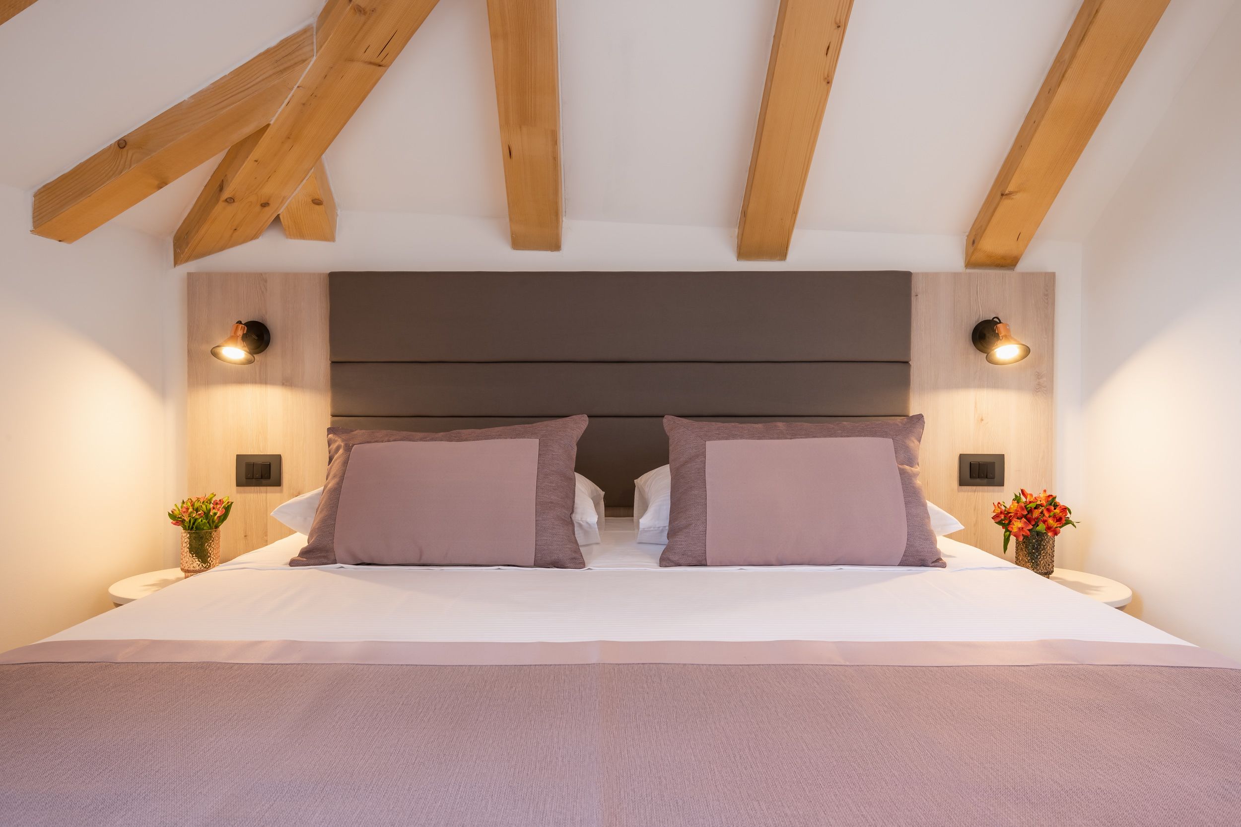 Un dormitorio con una cama grande, vigas de madera, dos luces fijadas a la pared y una mesita de noche con un florero.