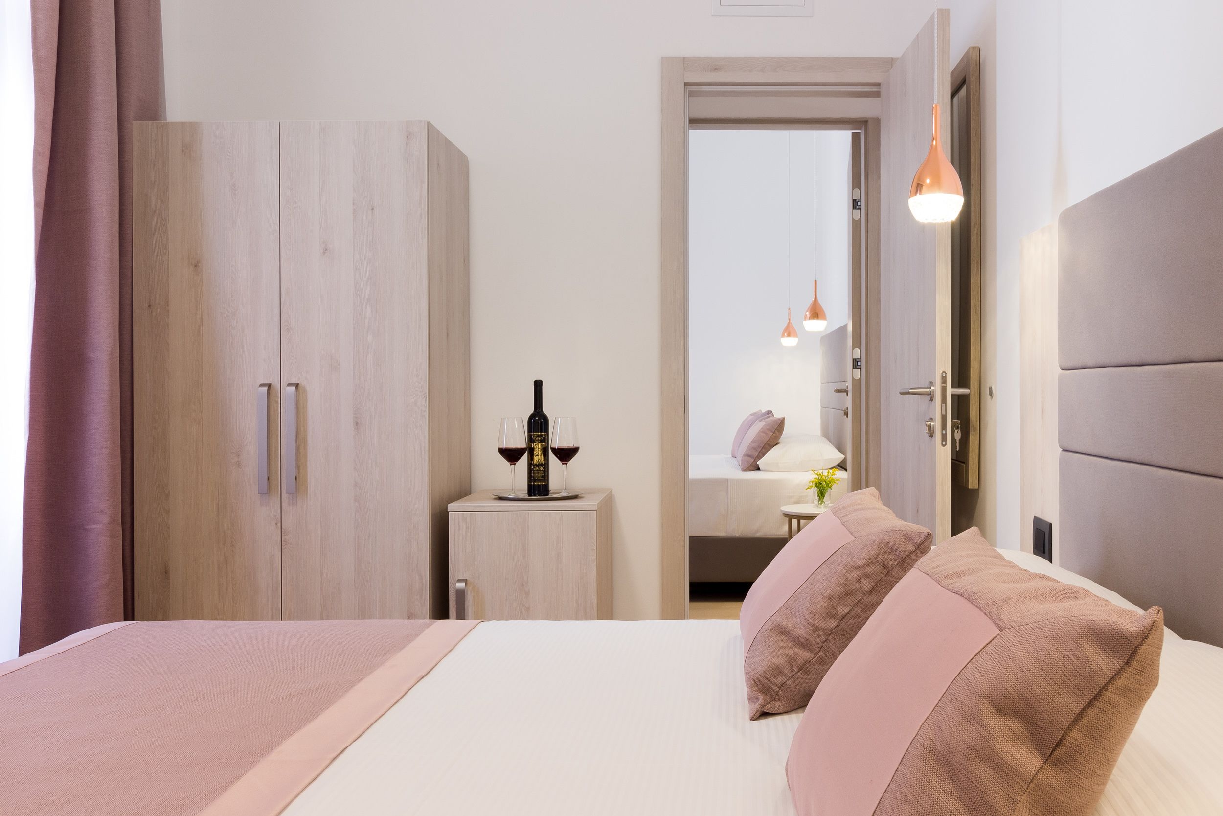 A hotel room with a bed, a wardrobe, a small table with a bottle and glasses, and a door leading to another room.