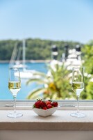 Un bol de fresas y dos vasos de vino blanco sobre un alféizar con vistas a barcos y follaje verde.