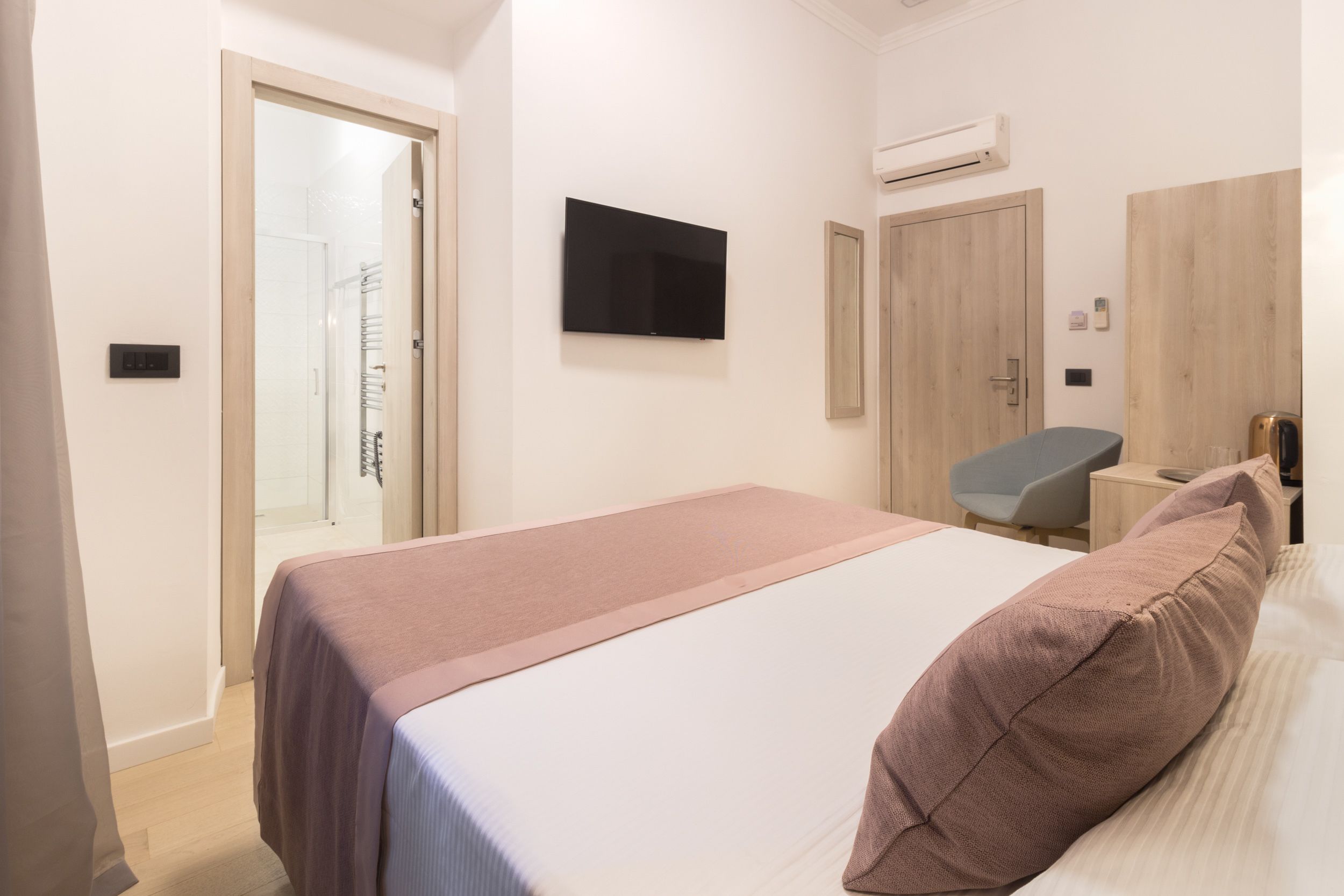 A hotel room with a bed, a TV mounted on the wall, a wooden wardrobe, a chair, and a small table.