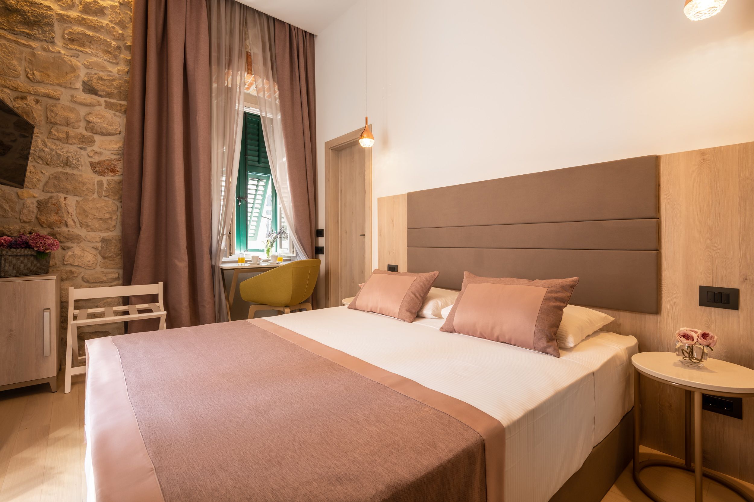A neatly arranged bedroom with a large bed, a small table, a chair, and a stone wall.