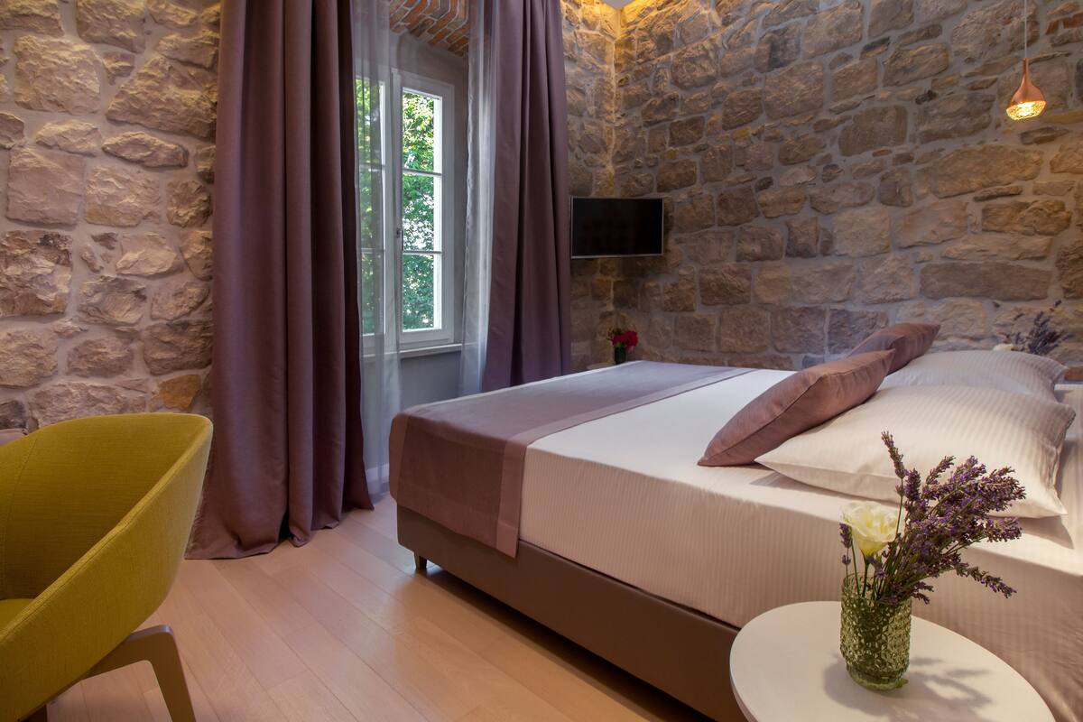 A bedroom with a stone wall, a bed with a white and brown bedspread, a yellow chair, and a small table with a vase of flowers.