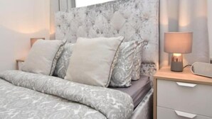 A neatly made bed with a patterned bedspread, a headboard with a textured design, and a bedside lamp on a wooden nightstand. - Cozy Apartment in Camden Town (London)