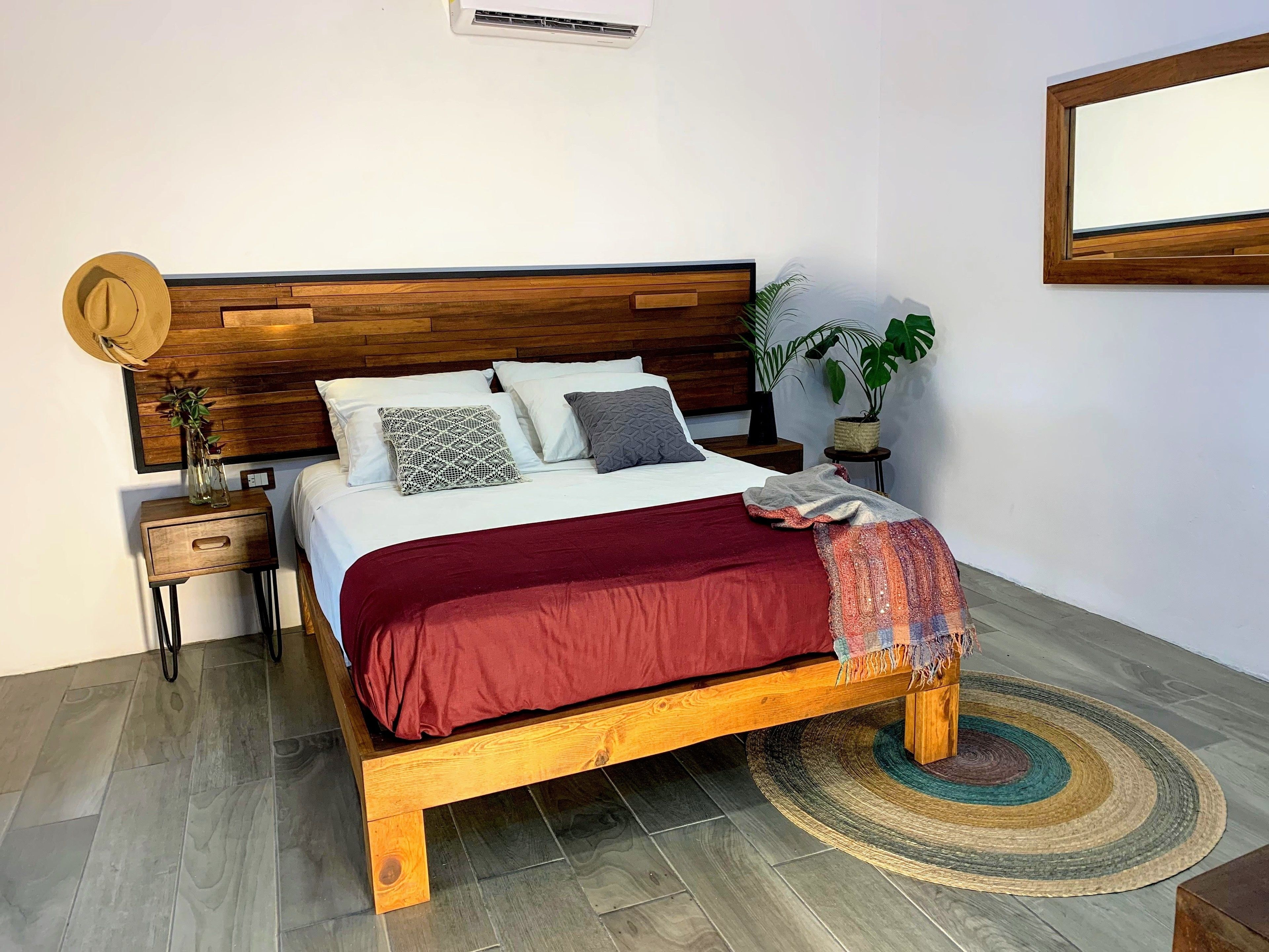 Quarto com cama de madeira, roupa de cama branca, manta vermelha, uma pequena mesa de cabeceira com uma planta e tapete redondo de fibra.