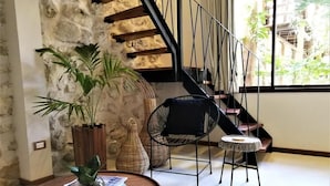 A cozy living area with a stone wall, a wooden table with a vase of plants, a wicker chair, and a view of a staircase leading to a balcony with plants. - Hotel Boutique TerraNova (Tulum)
