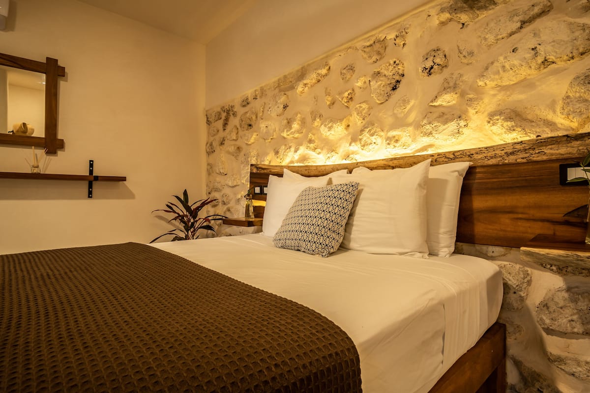 Un dormitorio con pared de piedra, cabecera de madera y cama con ropa de cama blanca y almohadas estampadas.