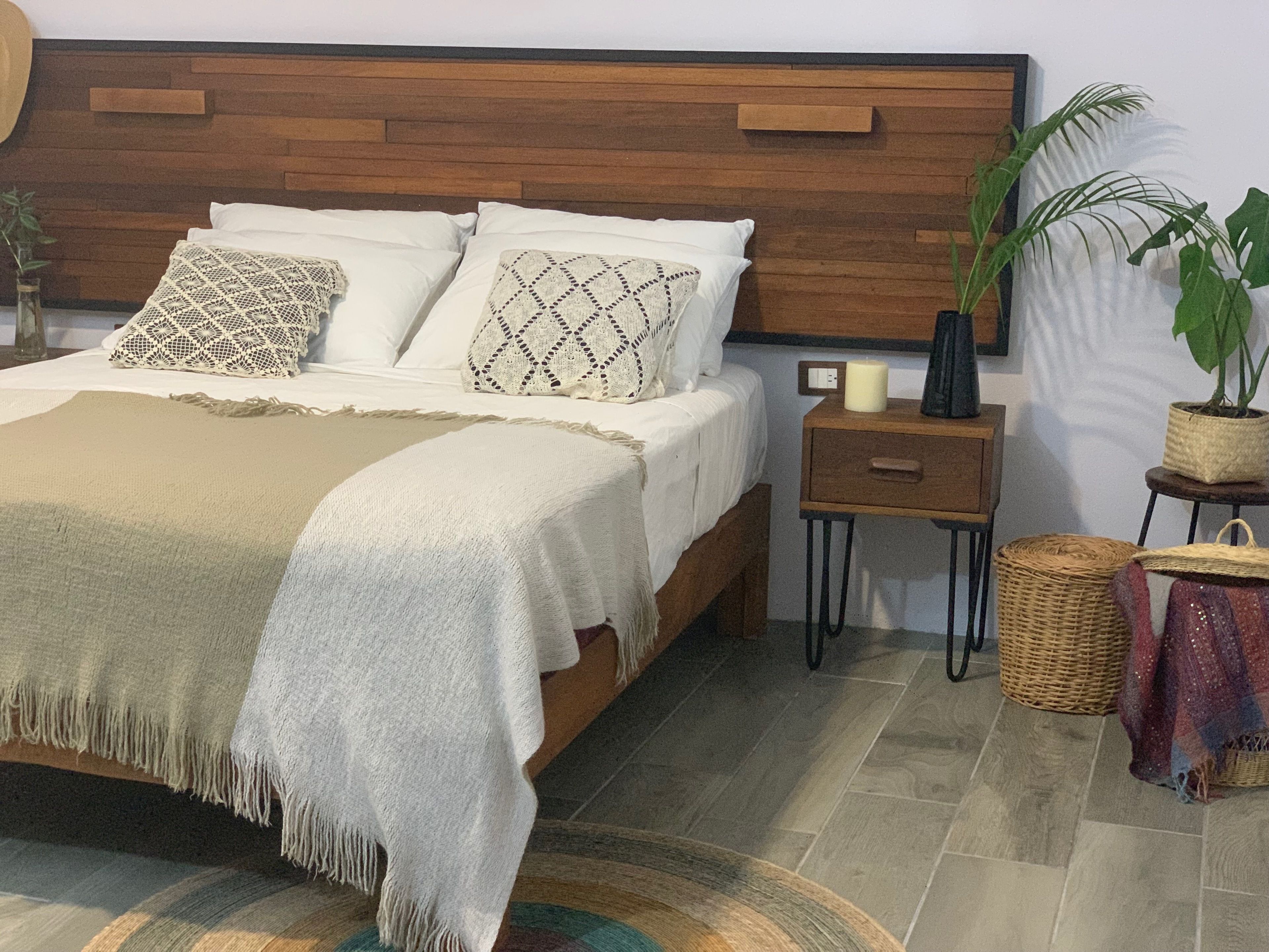 A bedroom with a wooden headboard, a bed with white linens, a beige throw blanket, a bedside table with a candle, and a potted plant.
