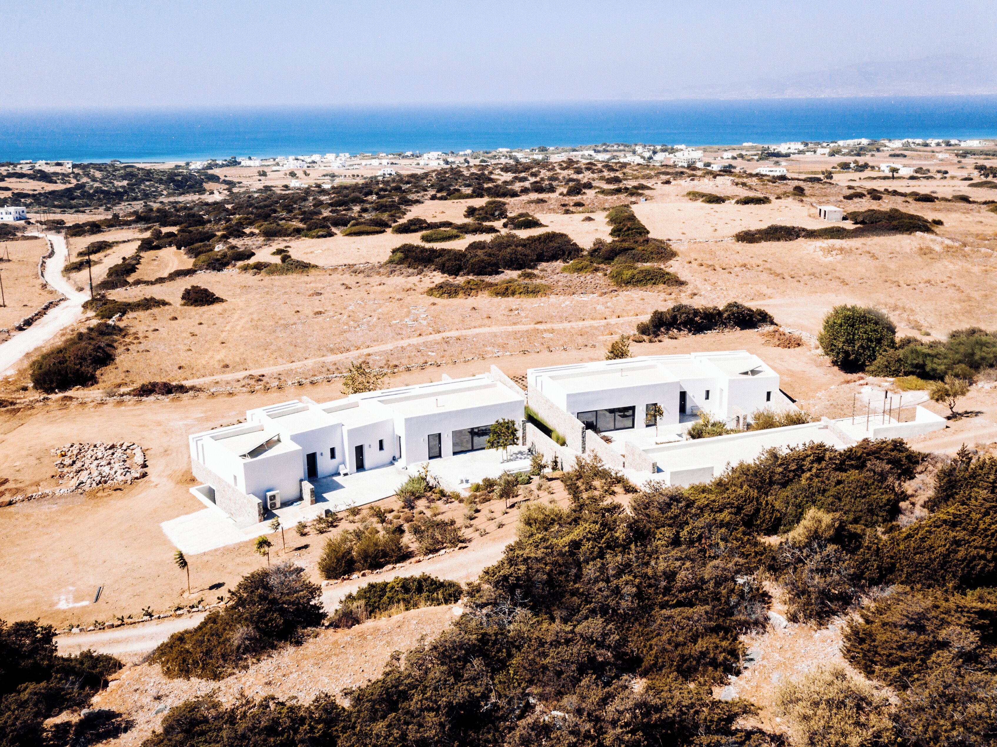 A modern white building with a flat roof, surrounded by sparse vegetation and a clear view of the sea in the background.