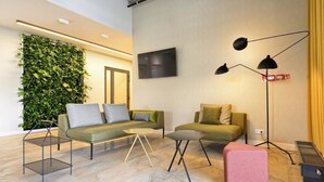 A modern living room with a green sofa, a small table, a pink ottoman, a floor lamp, and a green wall with plants. - Arche Hotel Poloneza (Warsaw)