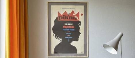 Une chambre d’hôtel moderne avec un canapé gris, des coussins jaunes et roses, une affiche encadrée au mur, une lampe sur pied et une petite table sur laquelle se trouvent un livre et une tasse.