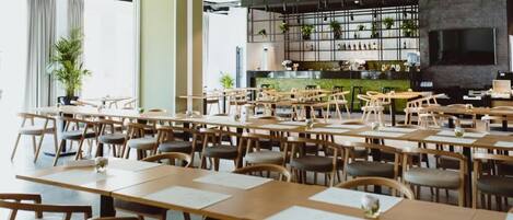 A spacious restaurant with multiple wooden tables and chairs, a bar area with shelves of bottles, and a large TV screen.