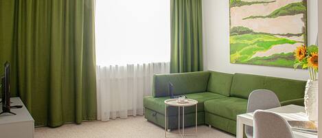 Een moderne woonkamer met een groene bank, een klein bijzettafeltje, een wit bureau en een stoel. Er hangt een schilderij aan de muur en er is een raam met dunne gordijnen.