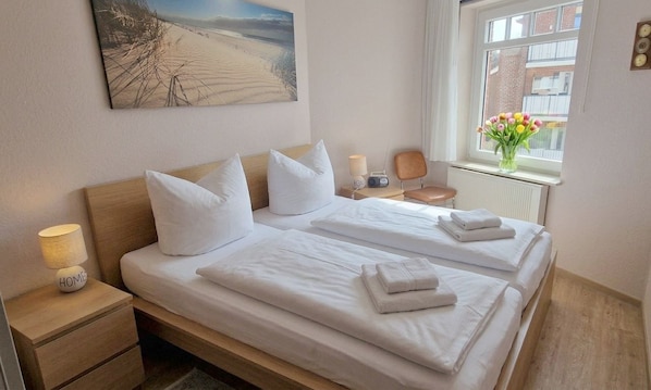 Un lit double avec du linge de lit blanc, une table de chevet avec une lampe, une chaise, une fenêtre ornée de fleurs et un tableau au mur.