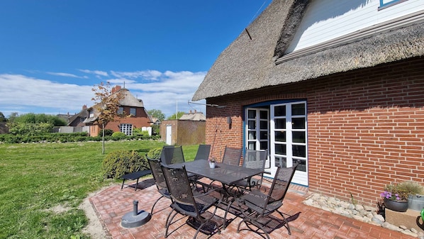 A patio with a table and chairs, a brick building with a thatched roof, and a grassy yard.