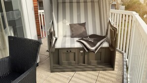 Ein Balkon mit gestreiftem Baldachin, einem Korbstuhl und einer gepolsterten Sitzecke mit Kissen und Decke.