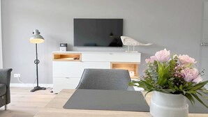 Ein modernes Wohnzimmer mit Flachbildfernseher, Stehlampe, grauem Sofa, grauem Sessel, einem kleinen Tisch und einer Vase mit Blumen.