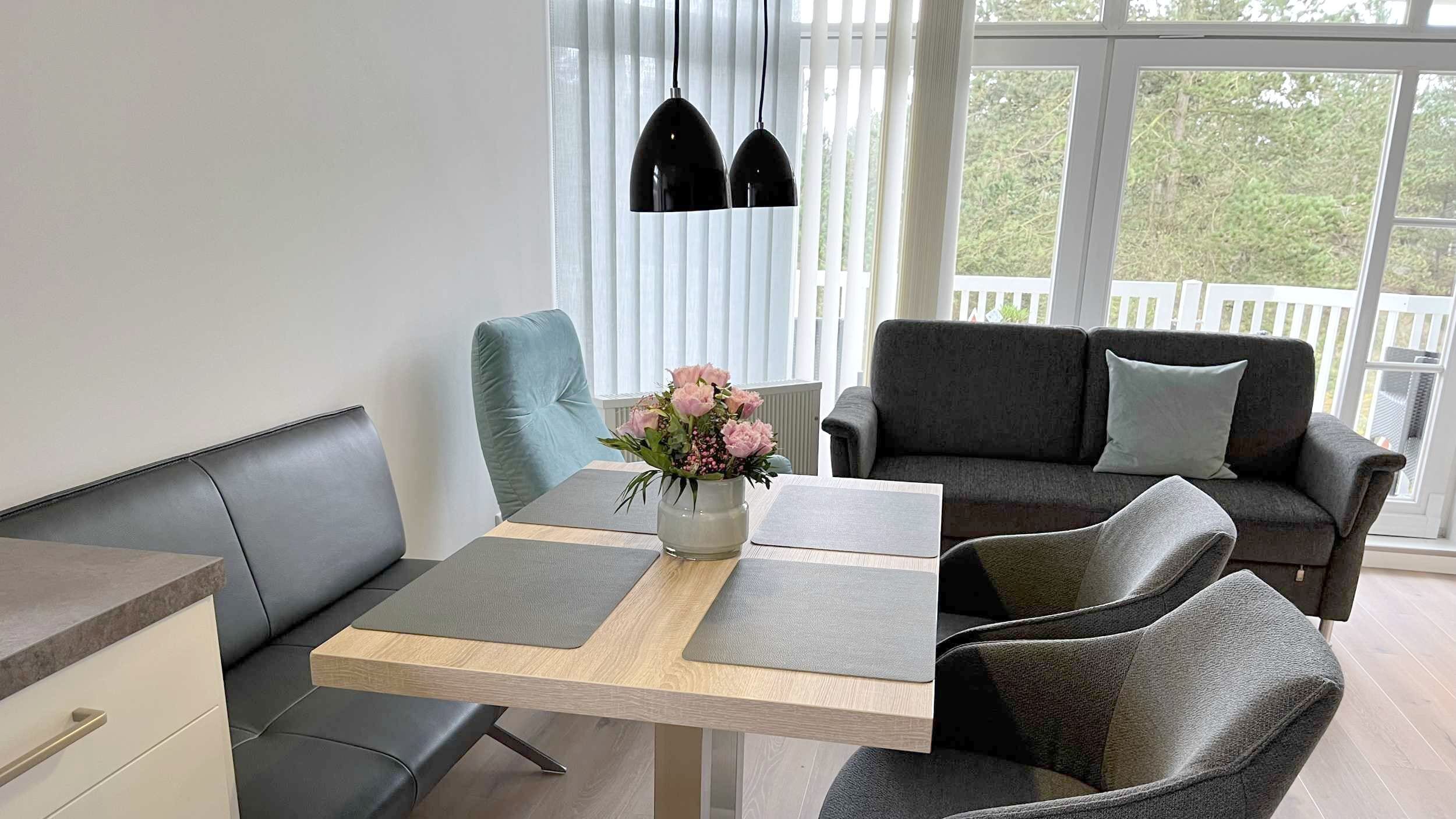 A modern living room with a dining table, grey sofa, and a vase with flowers.