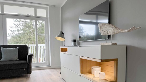 Ein modernes Wohnzimmer mit großem Fenster, einer Couch, einem an der Wand montierten Fernseher und einem Kamin mit Kerzen.