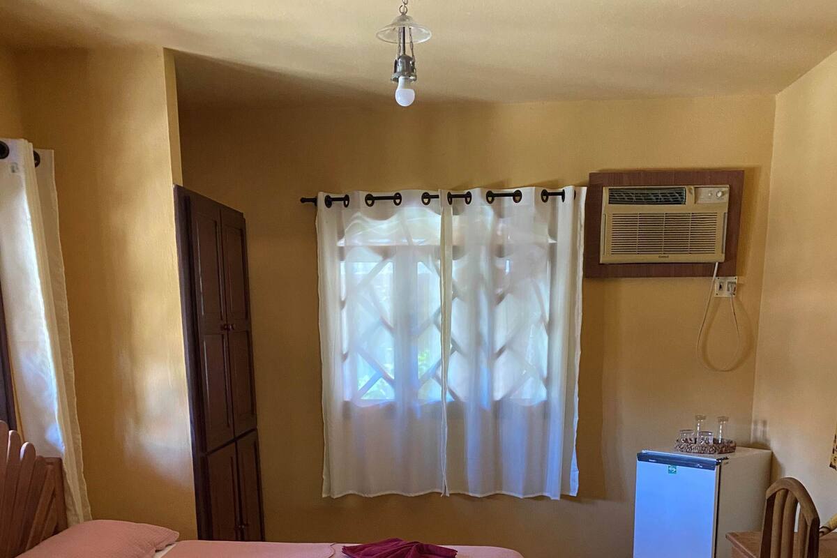 Quarto com cama, janela com cortinas, ar condicionado, pequena mesa e cadeira.