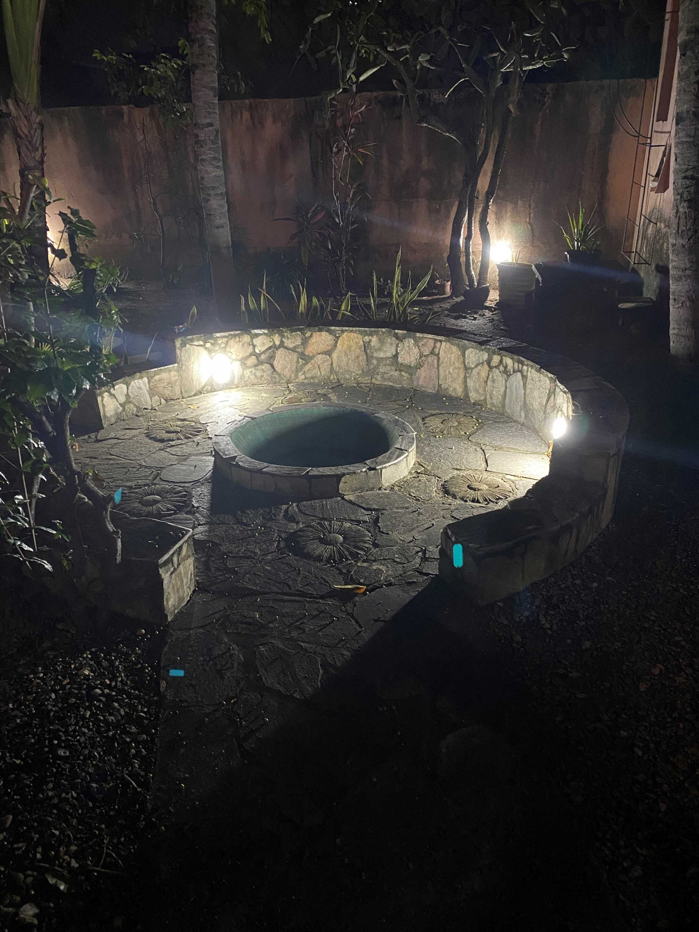 Un hogar de piedra con zona de asientos circular, rodeado de plantas y árboles.