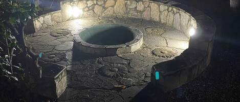 Un hogar de piedra con zona de asientos circular, rodeado de plantas y árboles.