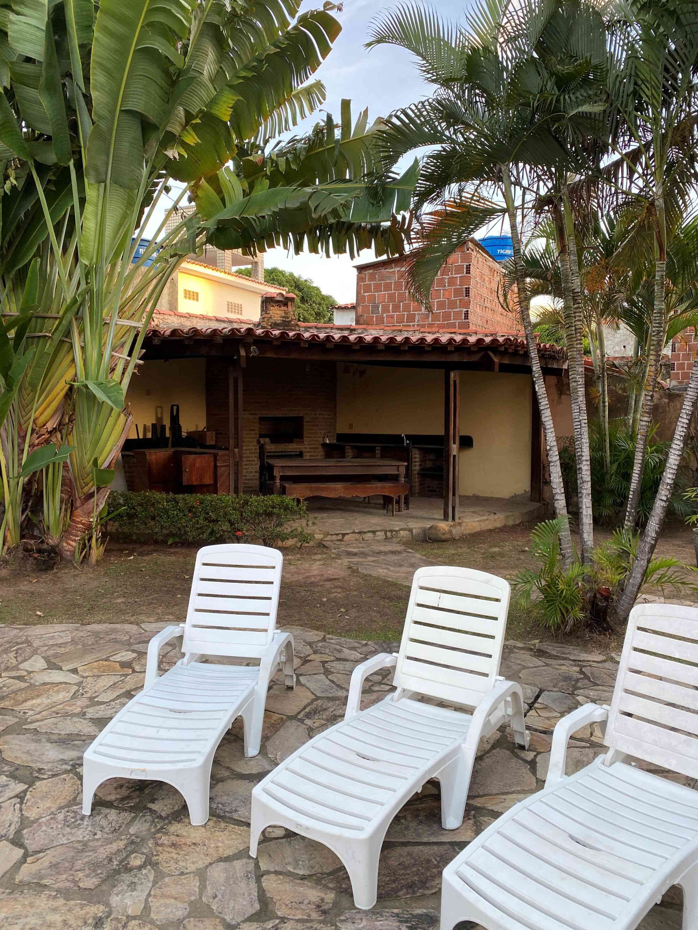 A patio with three white lounge chairs, a stone floor, and a brick building with a tiled roof in the background.